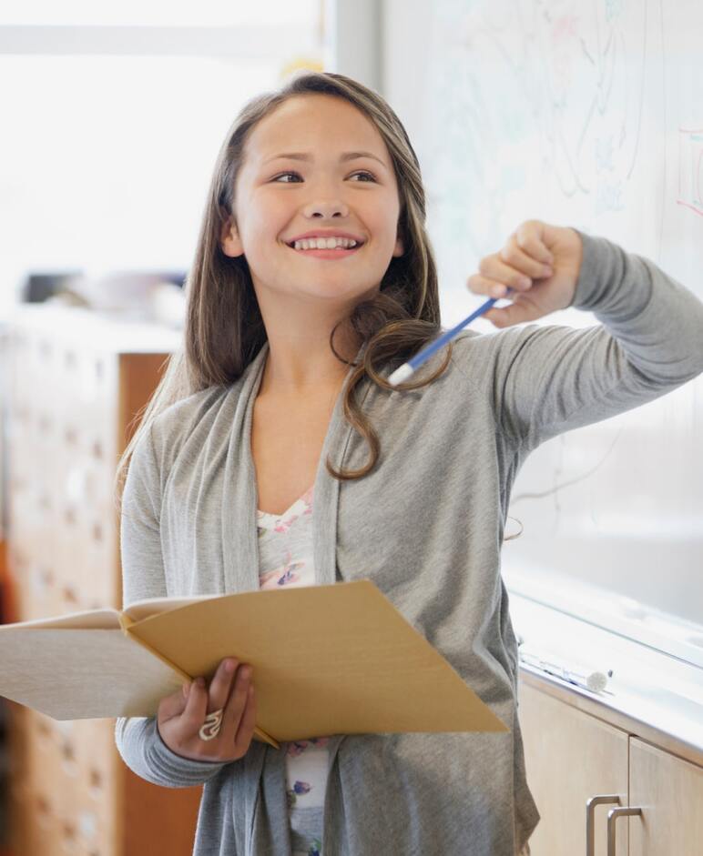 A teacher holding a folder and pointing to a whiteboard with SEL strategies in a classroom.”